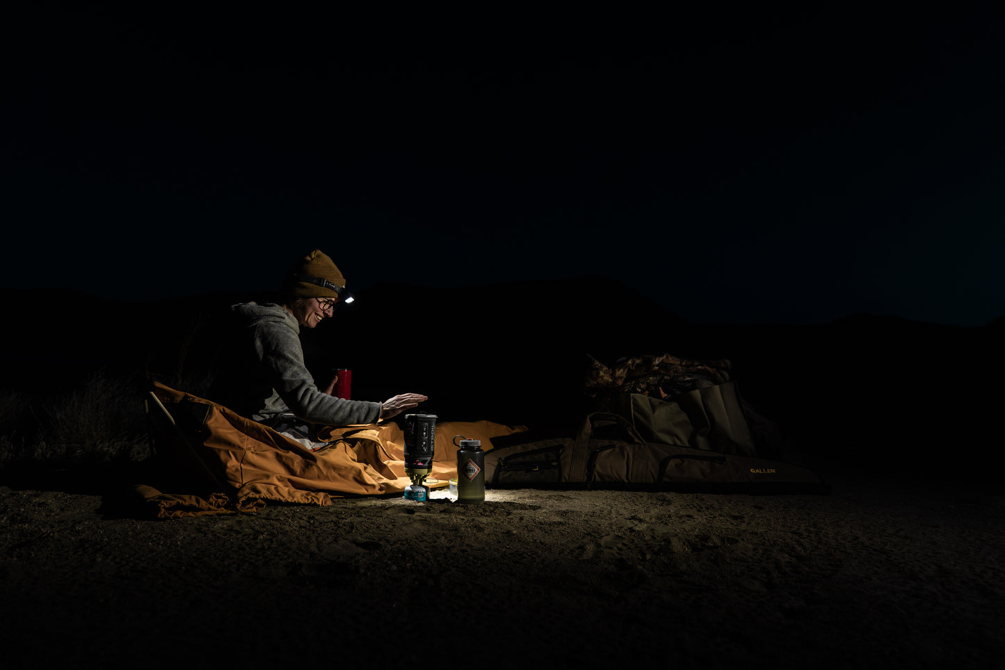 Woman sitting up in bedroll with headlamp in the dark next to jet boil.