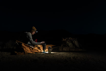 Woman sitting up in bedroll with headlamp in the dark next to jet boil.