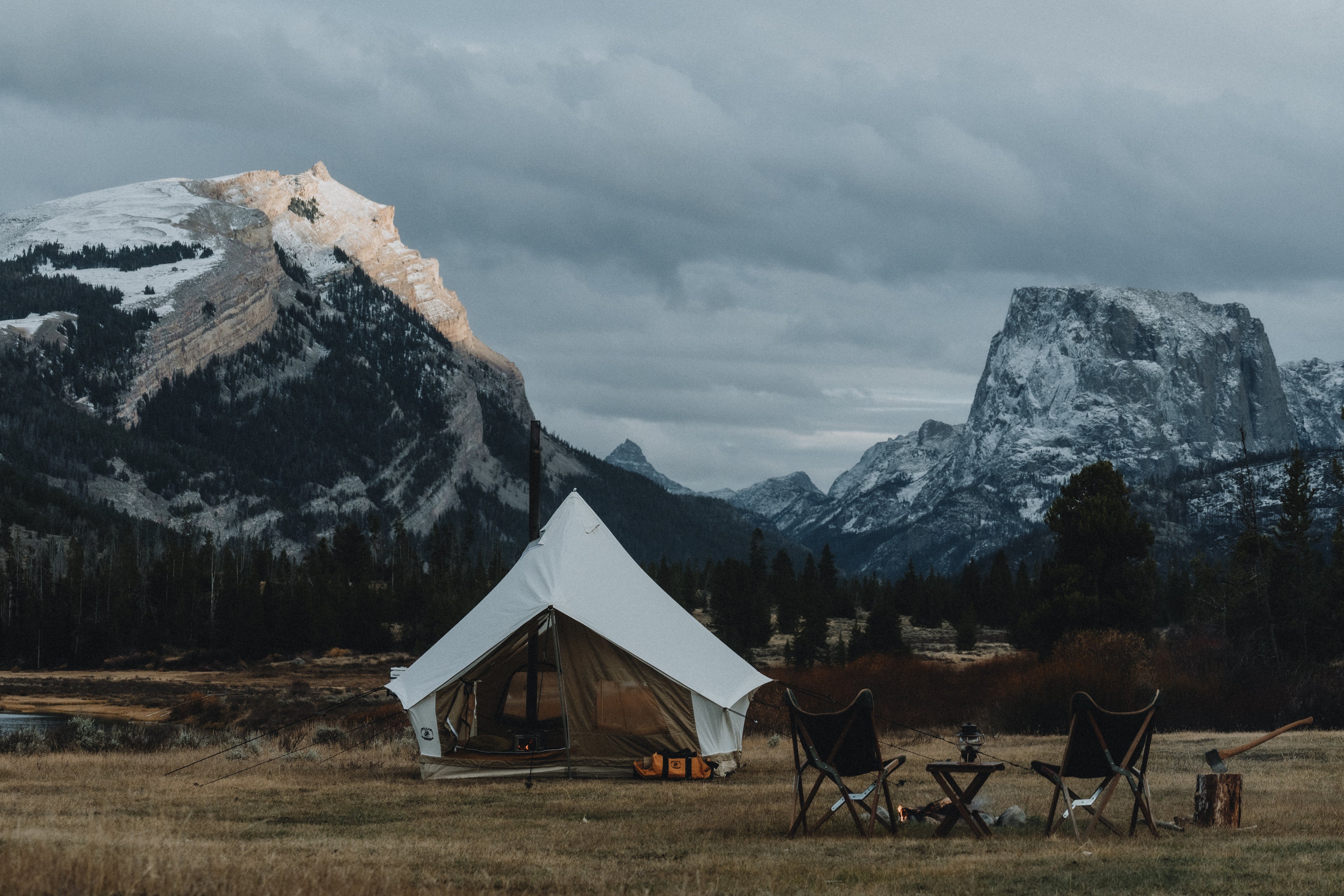 Square center pole canvas tent with a  woodstove in the WindRiver Range snow capped mountains in the distance. Two camp chairs with a chair and table around the fire out in front.